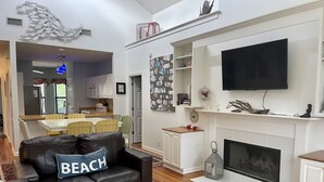 A modern living room with a fireplace, a black leather sofa with a 'BEACH' pillow, a dining area with yellow chairs, and a kitchen in the background. - Charming Beach House by Deer Lake Pool & Pond (Fripp Island)
