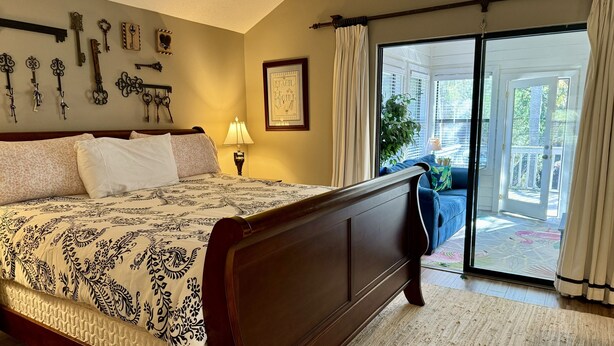 A bedroom with a large bed, a nightstand with a lamp, a wall-mounted key collection, and a sliding glass door leading to a balcony. - Charming Beach House by Deer Lake Pool & Pond (Fripp Island)