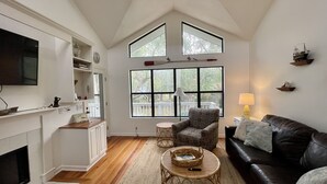 A spacious living room with a fireplace, a large window, a black leather sofa, and a patterned armchair. - Charming Beach House by Deer Lake Pool & Pond (Fripp Island)