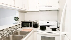 Cozinha compacta com armários brancos, pia de aço inoxidável, micro-ondas, fogão e bancada pequena com tábua de corte.