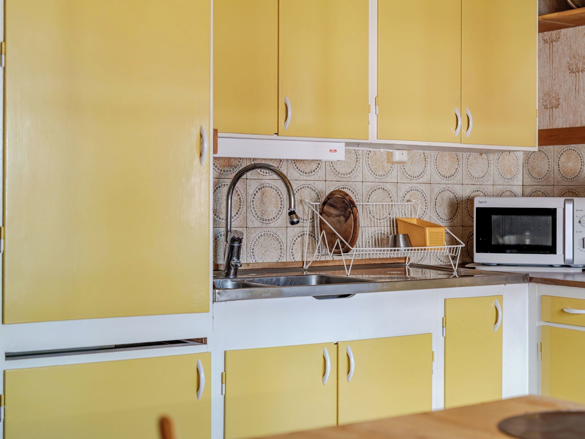 Een keuken met gele kastjes, een roestvrijstalen spoelbak en een witte magnetron.