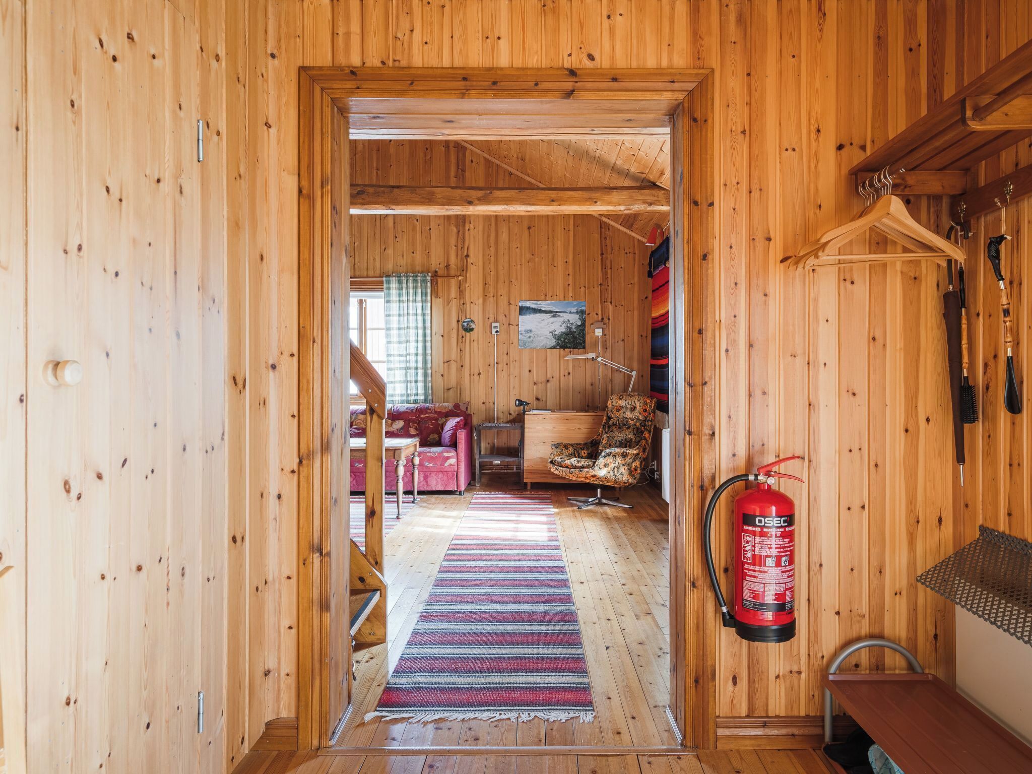 En träinredd interiör med en hall som leder till ett vardagsrum med en soffa, en fåtölj, en öppen spis och en brandsläckare.