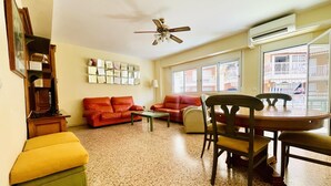 A living room with a red sofa, a yellow ottoman, a wooden coffee table, and a dining table with chairs. There is a ceiling fan, a wall-mounted air conditioner, and a window with a view of a balcony.