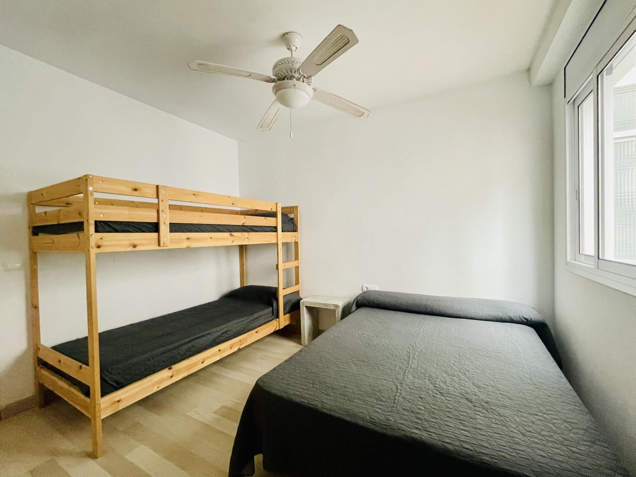 A dormitory room with a bunk bed, a single bed, a small table, and a ceiling fan.