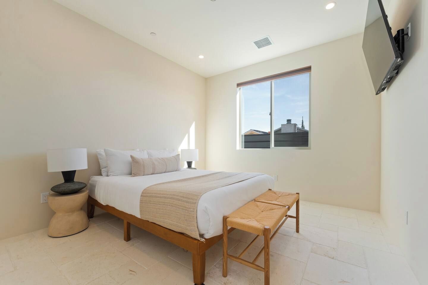 Quarto com cama, mesas de cabeceira, um banco, janela e televisão na parede.