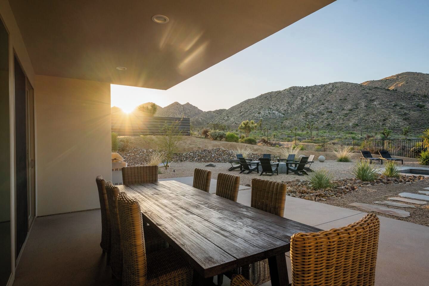 Une terrasse extérieure avec une table en bois et des chaises en osier, offrant une vue sur un paysage désertique et des montagnes.
