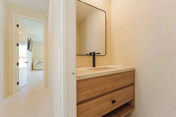 A bathroom with a wooden vanity, a mirror, and a white door leading to a living area.