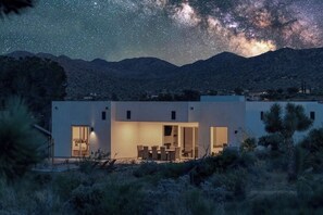 A modern house with a dining area and a large window, set against a mountainous backdrop with a starry sky.