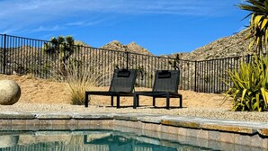 A swimming pool with two lounge chairs, a stone wall, and a desert landscape in the background.
