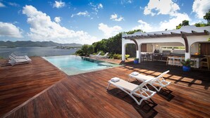 Eine Holzterrasse mit Pool, Liegestühlen und Grillplatz mit Blick auf ein Gewässer.