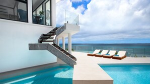 Een modern zwembadgedeelte met een glazen balustrade, een trap naar een balkon en loungestoelen met uitzicht op de oceaan.