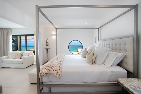 Un dormitorio moderno con cama con dosel, vista al mar a través de un ventanal circular y una decoración en tonos blancos.