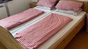 Une chambre avec un lit simple, un cadre en bois, une couvre-lit à carreaux, une table de chevet avec une lampe et un tableau représentant un paysage montagneux.