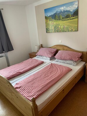 A single-bed room with a wooden frame, a checkered bedspread, a bedside table with a lamp, and a painting of a mountainous landscape.
