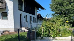 Une allée pavée mène à un bâtiment blanc doté d’un toit sombre, entouré de verdure et délimité par une clôture en métal.