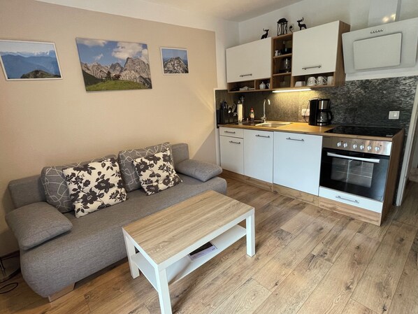 A compact living space with a sofa, a small table, a kitchenette with a stove, and wall-mounted cabinets. - Ferienwohnung für 2 Erwachsene und 1 Kind, 1 Separater Schlafraum, Terrasse, 40 m² (Inzell)