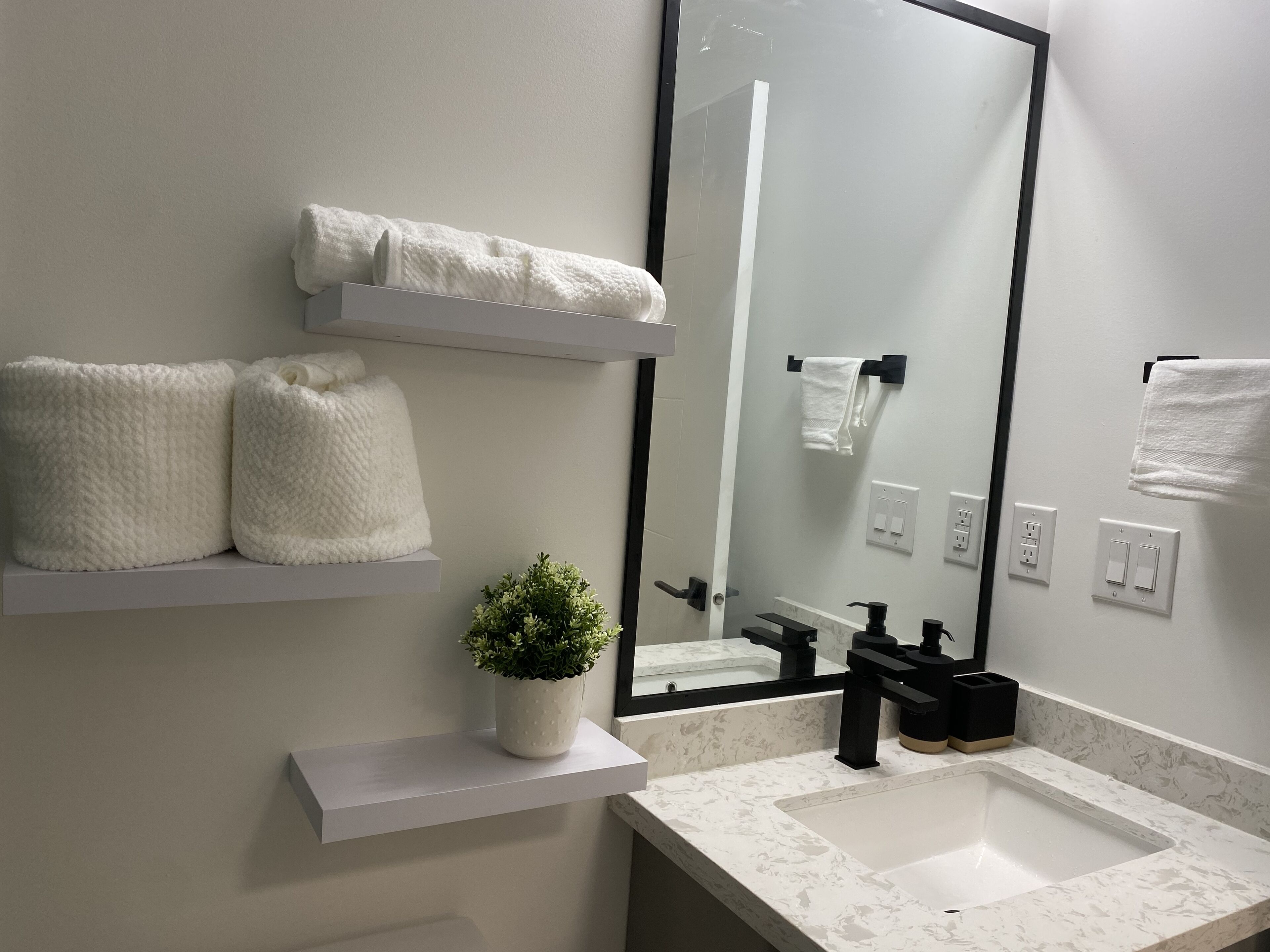 A bathroom with a white sink, black faucet, and a mirror. There are two white towels on shelves and a small potted plant on the countertop.