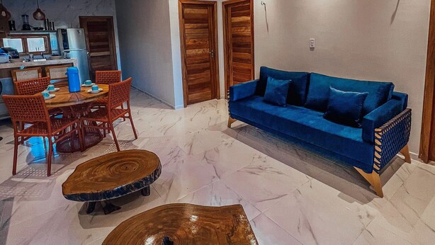 A modern living room with a blue sofa, wooden dining table, and chairs, a kitchen area with appliances, and a ceiling with red lighting. - Chill House (Cruz)