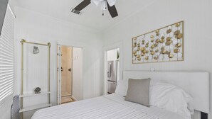 Une chambre dotée d’un grand lit, d’un ventilateur de plafond, d’une œuvre d’art accrochée au mur et d’une porte donnant accès à une salle de bain.