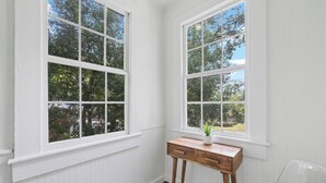 Une pièce aux murs recouverts de panneaux blancs, un bureau en bois doté de tiroirs et deux grandes fenêtres donnant sur des arbres.
