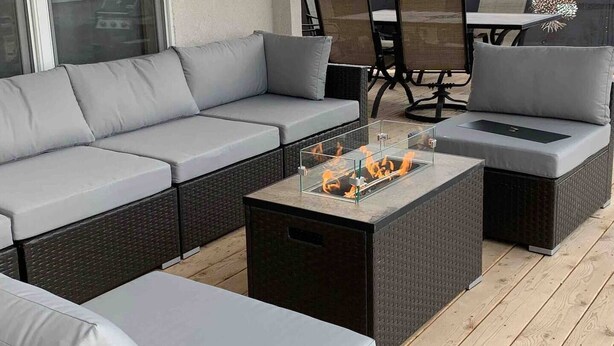 Un área de estar moderna al aire libre con un sofá gris, una mesa de centro de vidrio con chimenea integrada y una mesa de mimbre oscuro.