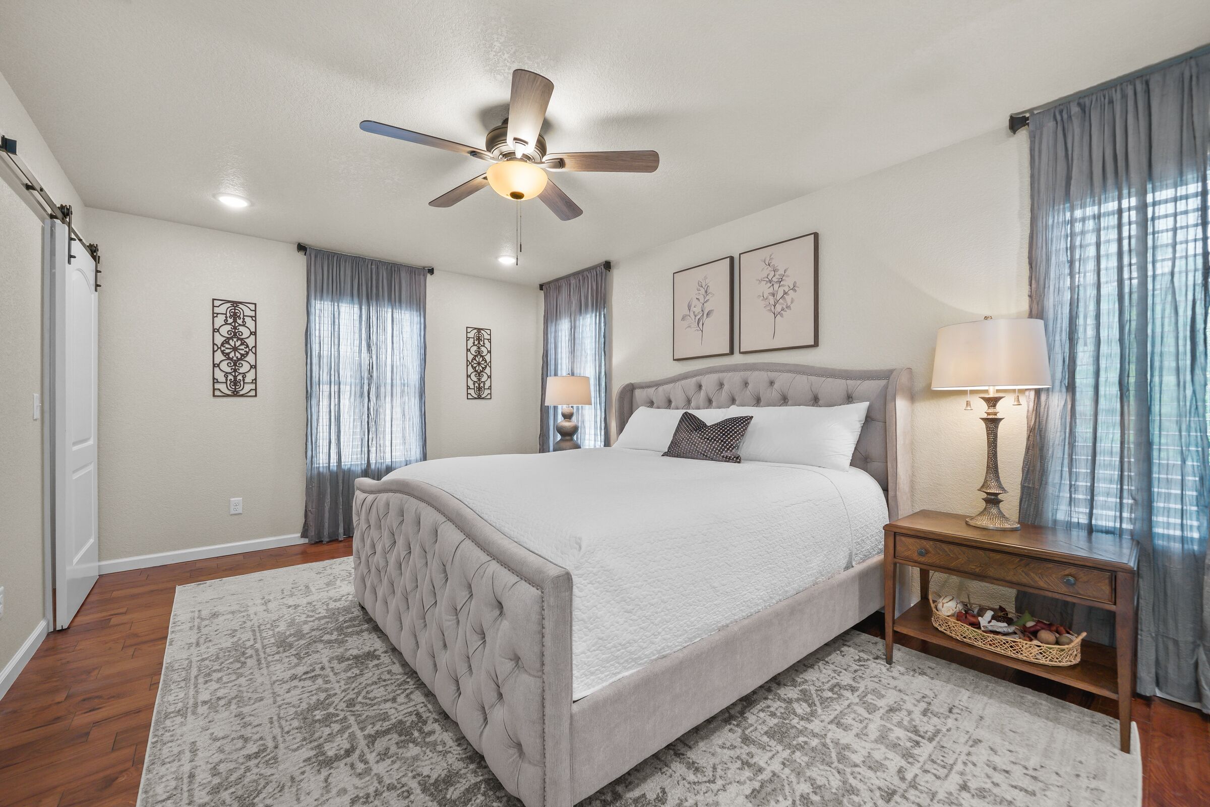 A bedroom with a tufted headboard bed, a ceiling fan, a bedside table with a lamp, and a decorative wall piece.