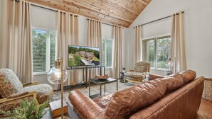 Una sala de estar con un sofá de cuero, un sillón estampado, una mesita de centro y un televisor que muestra un paisaje pintoresco.