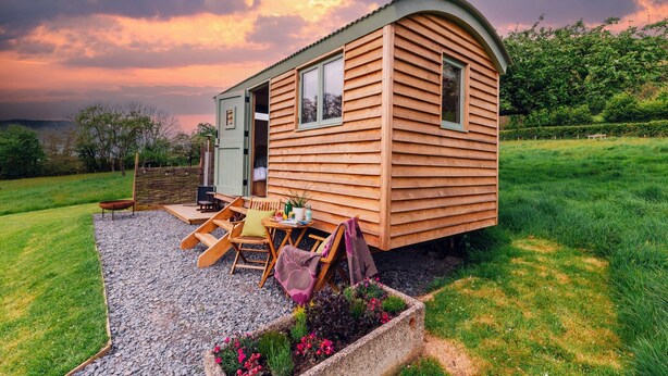 Una caba ña de madera con techo curvo, un patio con mobiliario exterior y un jardín lleno de flores.