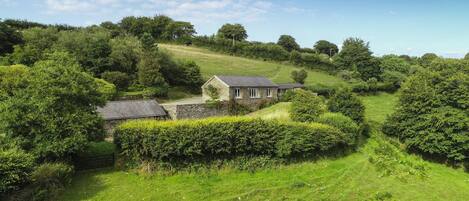A stone house with a sloped roof, surrounded by lush greenery and a clear sky.