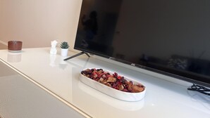 A flat-screen TV on a white stand, a small potted plant, a bowl of dried flowers, and a candle on a white surface.