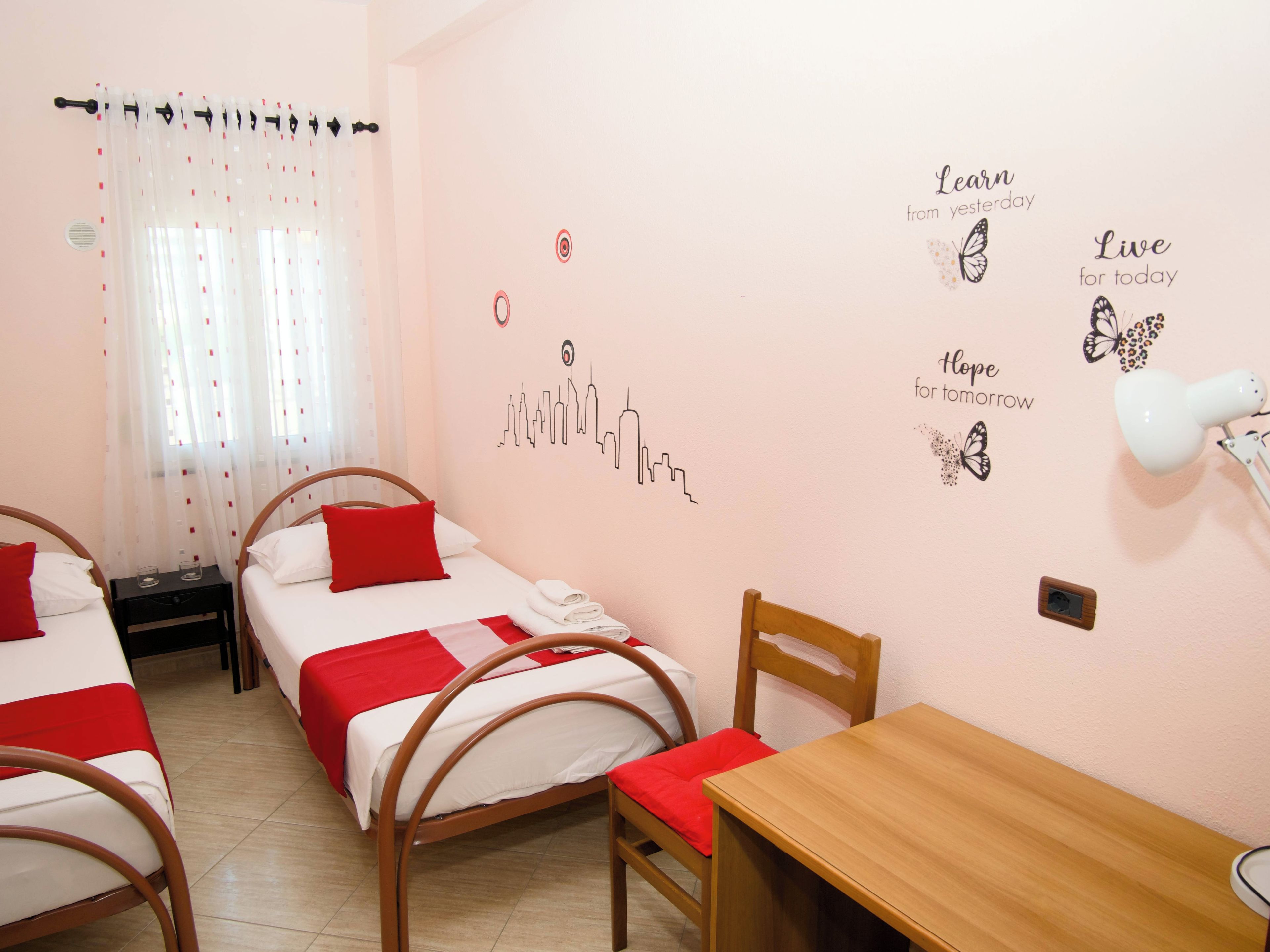 A hotel room with two single beds, a wooden table, a chair, a desk, and a window with sheer curtains.