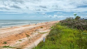 Une plage avec un rivage sablonneux, une zone herbeuse et un château d’eau visible au loin.