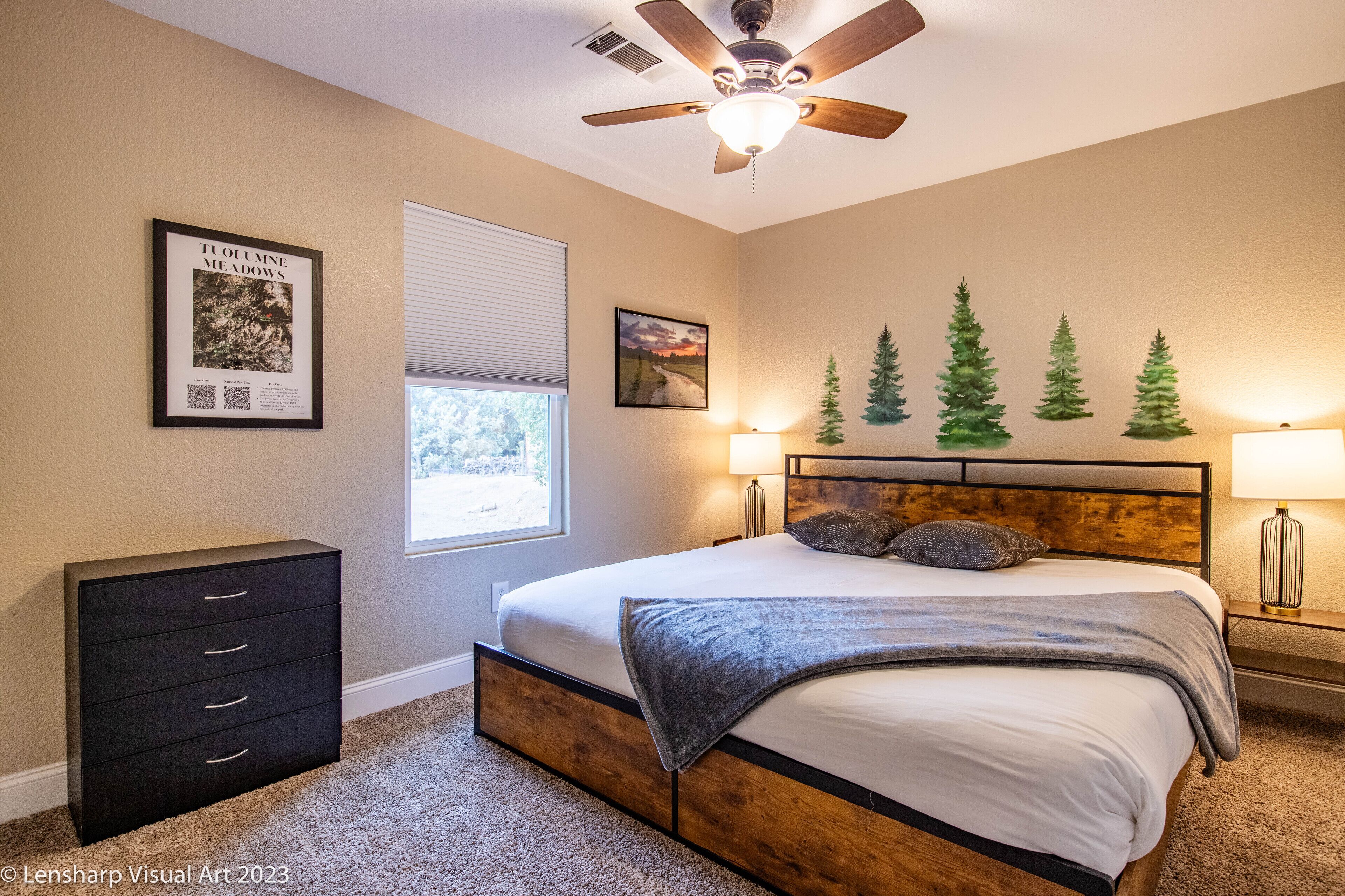 Une chambre à coucher comprenant un lit, un ventilateur de plafond, une commode et des éléments de décoration murale, notamment un tableau encadré et une fresque représentant des arbres.