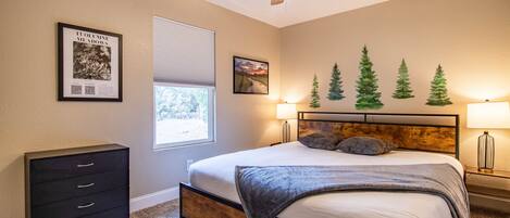Une chambre à coucher comprenant un lit, un ventilateur de plafond, une commode et des éléments de décoration murale, notamment un tableau encadré et une fresque représentant des arbres.