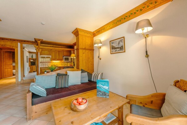 A cozy living area with a wooden sofa, a wooden table with a bowl of apples, and a book titled 'CHANONIS MONTBLANC'.