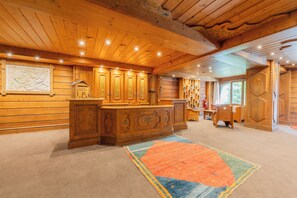 A wooden interior with a central wooden counter, bookshelves, and a large rug.