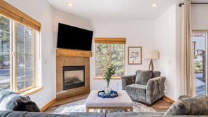 A living room with a fireplace, grey sofas, a flat-screen TV, and large windows. - Bright 3BD Kings Beach Townhome (Kings Beach)