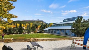Un edificio moderno con ventanas amplias, un patio empedrado con bancos y vistas a una ladera boscosa.