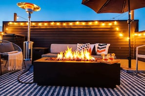 Un foyer avec des flammes, un canapé avec des coussins et des guirlandes lumineuses sur une terrasse en bois.