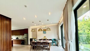A modern kitchen with wooden cabinets, a dining area with a table and chairs, and a large window with curtains. - Beach villa 3BR private swimming pool (Phu Quoc)