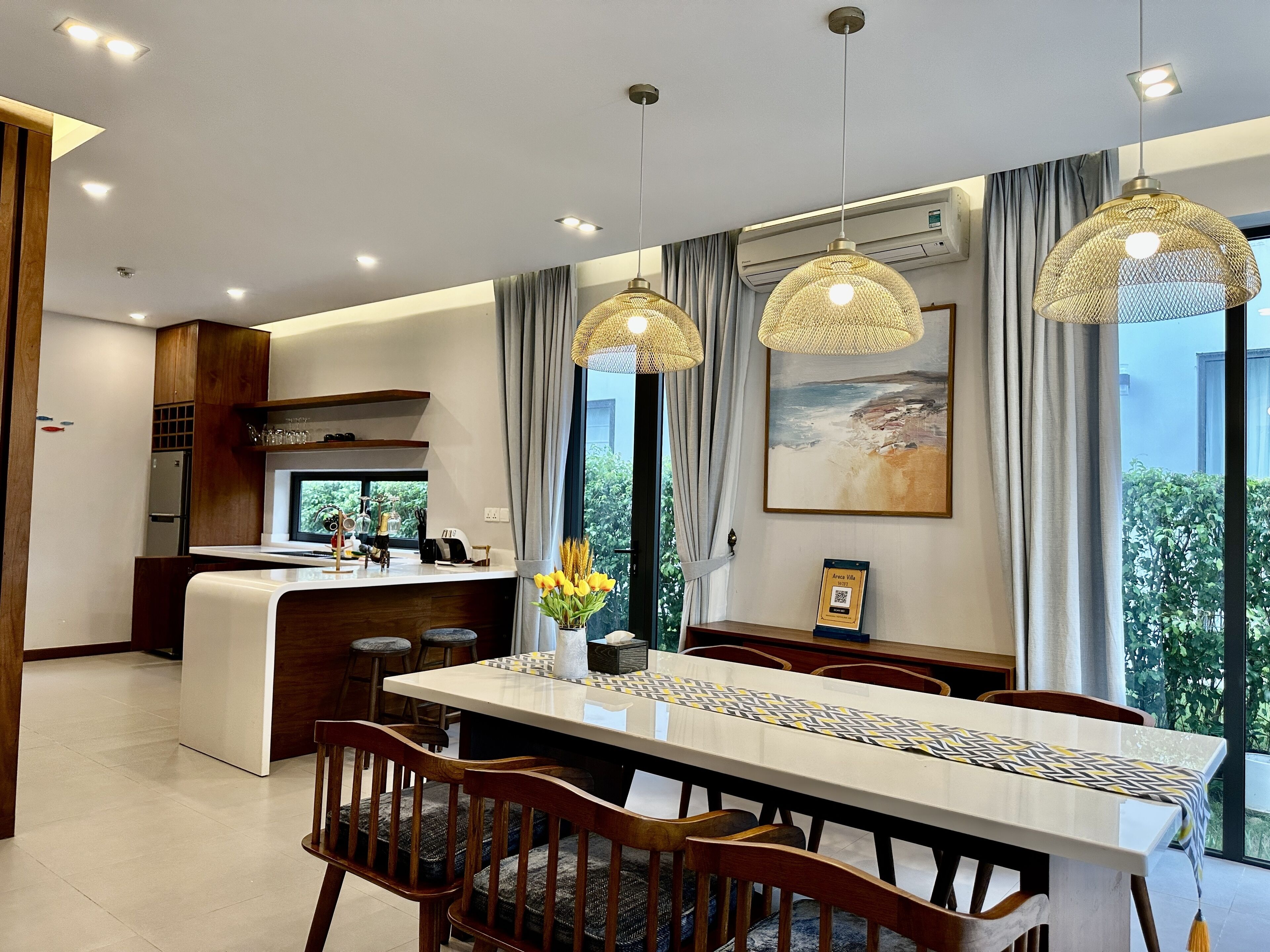 A modern kitchen with a central island, dining area with wooden chairs, pendant lights, and a view of greenery outside.