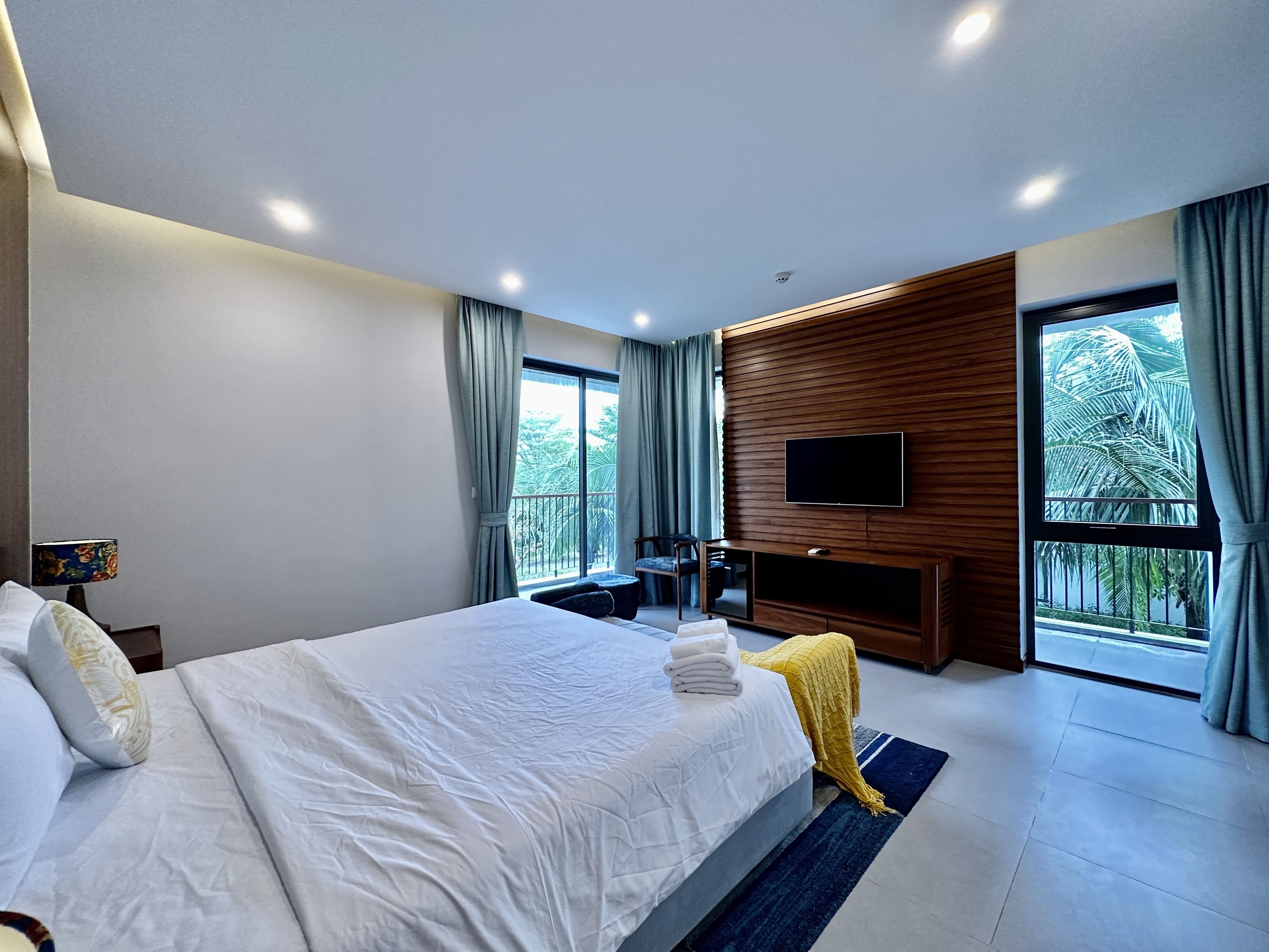 A modern hotel room with a large bed, a flat-screen TV mounted on the wall, a balcony with curtains, and a view of palm trees.