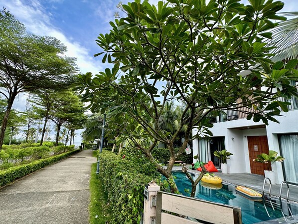 A pool area with a clear view of the pool, a yellow float, and a red chair. There is a pathway lined with greenery and a building with a white facade in the background.