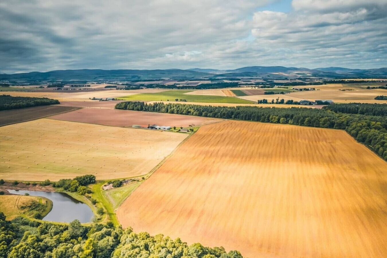 Ett flygbildsbild av ett lantligt landskap med åkrar, en liten sjö eller damm och avlägsna kullar.