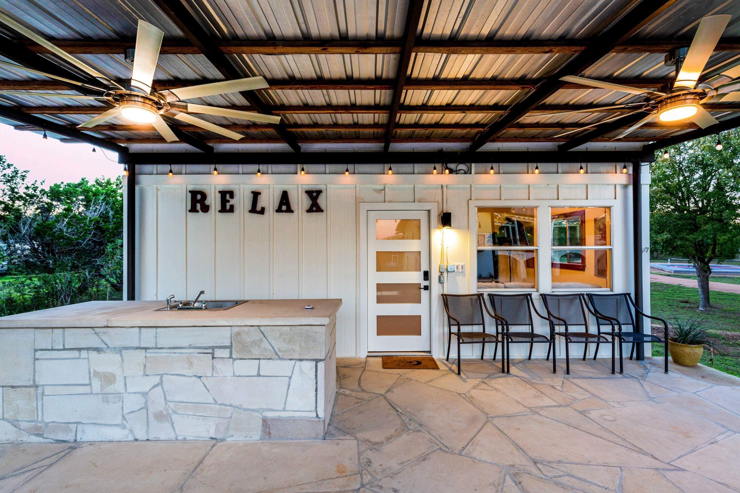 Un espace extérieur abrité avec un comptoir en pierre et des chaises, une pancarte indiquant « RELAX » et une vue sur la verdure environnante.