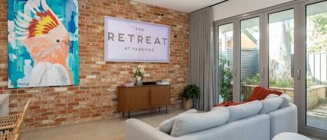 A modern living room with a large sofa, a coffee table, a painting of a bird, a sign reading 'The Retreat at Parkside,' and a glass door leading to an outdoor area.