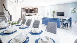 A modern dining area with a long table set for six, a kitchen in the background, and a living area with a blue sofa and a TV. - Champions Gate 8BR Villa-pool Game Room Theater (Davenport)