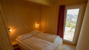 A bedroom with a wooden floor, a bed with two pillows, a nightstand, a wall-mounted light, and a window with a view of the outdoors. - Alpenchalet 341 by Interhome (Klippitztörl)