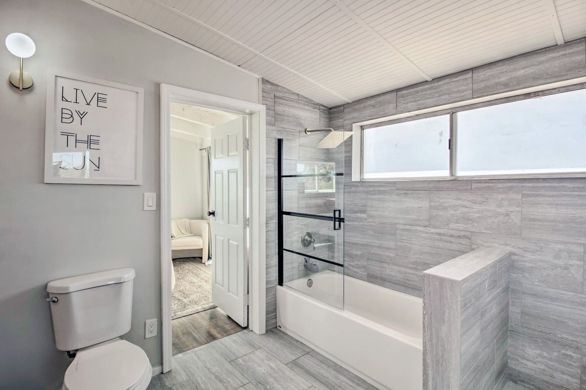 A modern bathroom with a glass-enclosed shower, a bathtub, and a toilet. There is a window providing natural light and a sign on the wall that reads "LIVE BY THE SUN".