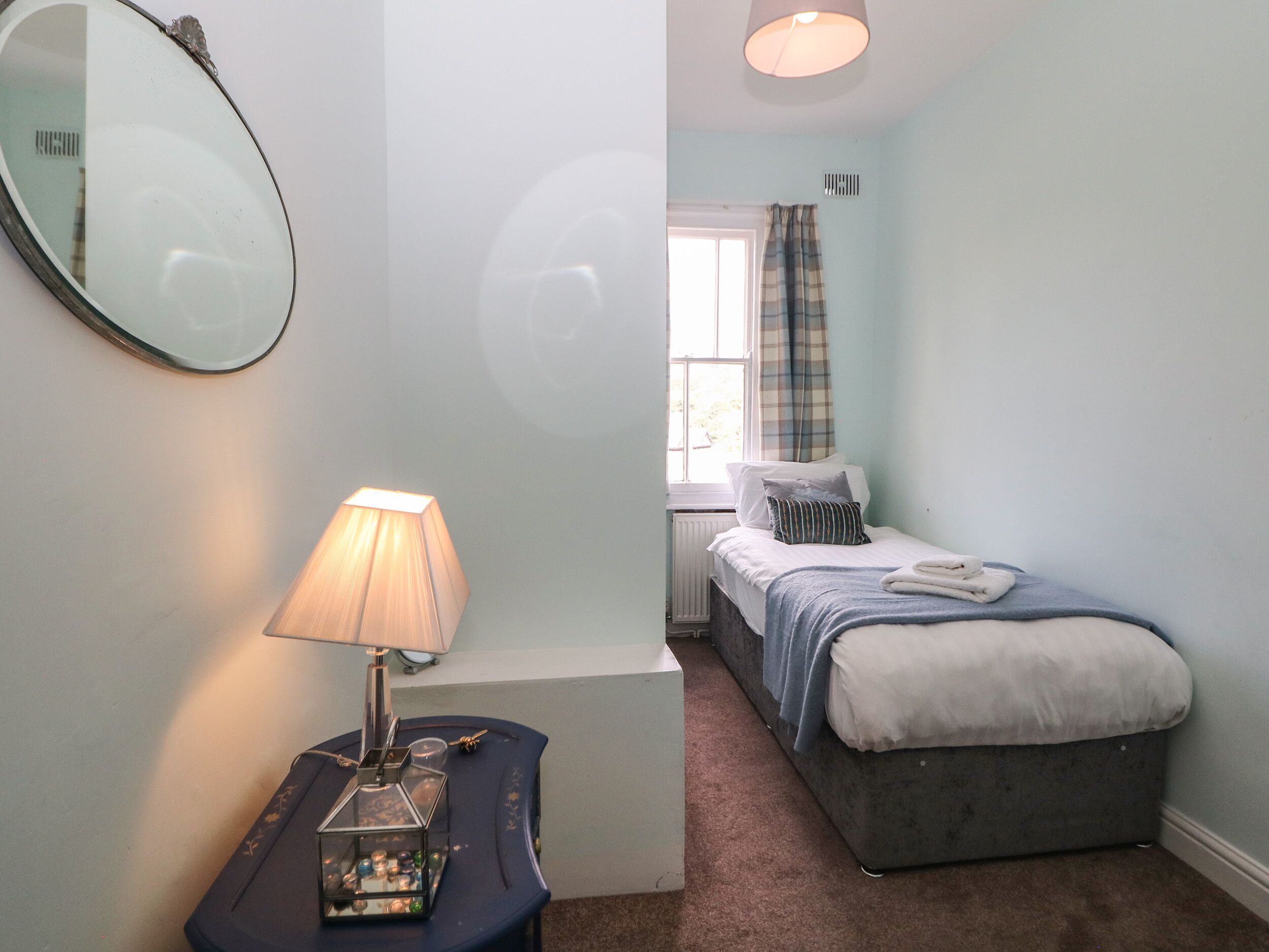 A bedroom with a bed, a bedside table with a lamp, a mirror, and a window with curtains.